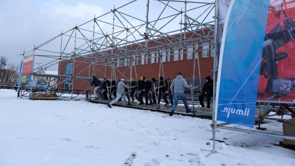 Eine Hollywood-Schaukel aus Gerüstmaterialien steht im Schnee. Einige Menschen geben der Schaukel Anschwung.