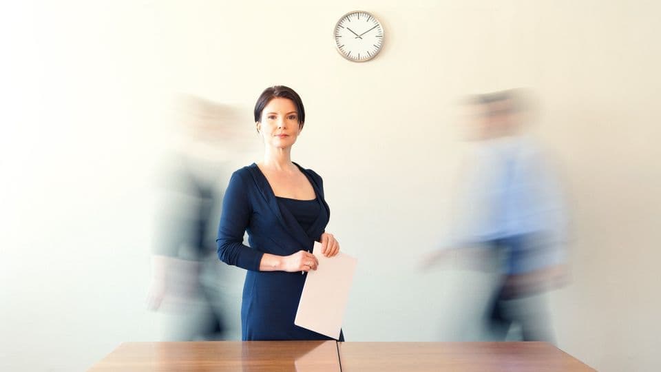 Frau steht im Buero an Konferenztisch, zwei Kollegen in Bewegung