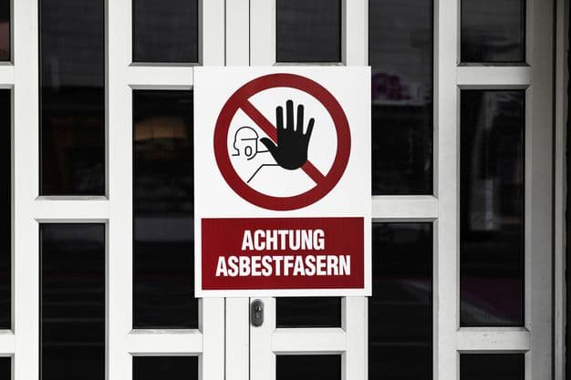 Weiße Haustür auf der ein Warnschild angebracht ist. Dort steht: "Achtung Asbestfasern".