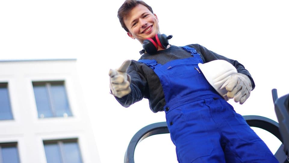 Junger Mann auf der Baustelle hat einen Bauhelm unter dem Arm und lächelt von oben in die Kamera