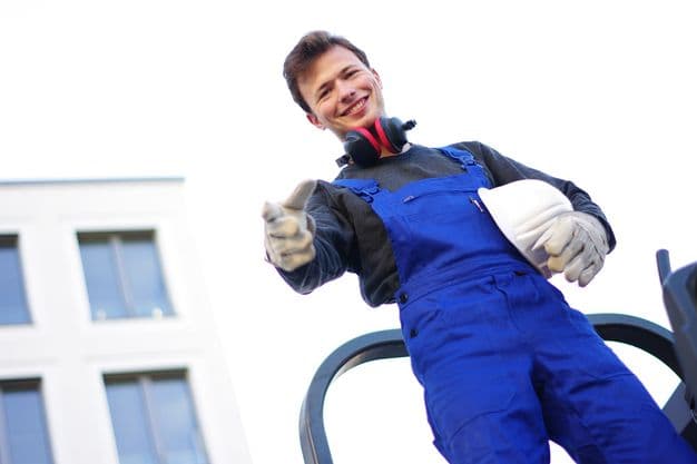 Junger Mann auf der Baustelle hat einen Bauhelm unter dem Arm und lächelt von oben in die Kamera