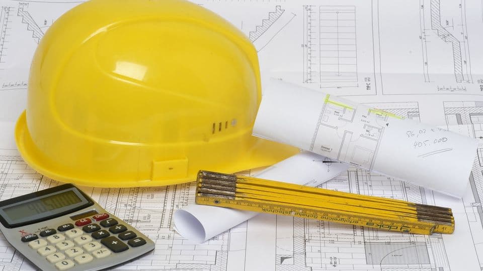 Bauplaene sowie in Helm, ein Zollstock und ein Taschenrechner liegen auf einem Tisch.