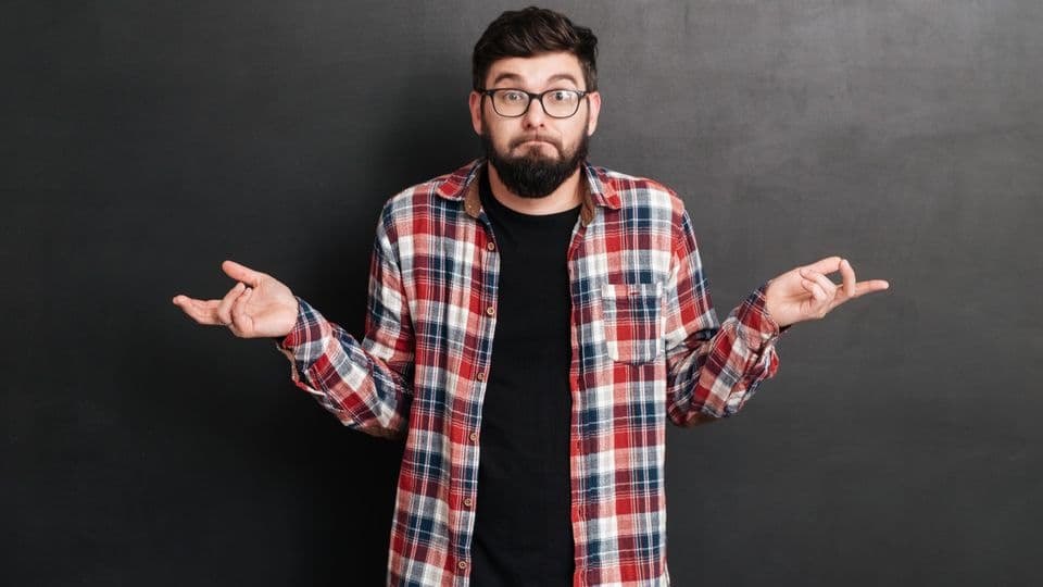 ahnungsloser studentConfused bearded young man in glasses and plaid shirt standing and shrugging his shoulders over grey background