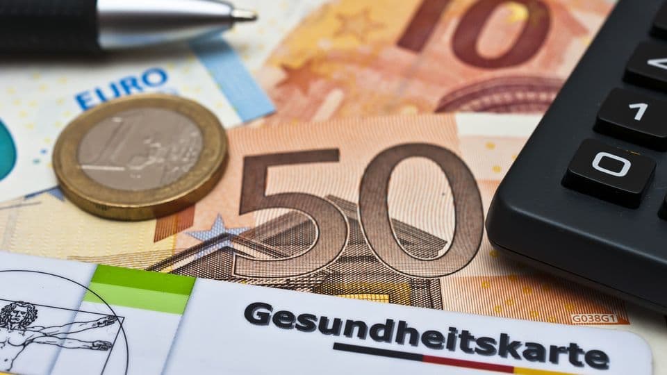 Eine Gesundheitskarte, ein Taschenrechner, ein Kugelschreiber und Geld liegen auf einem Schreibtisch.