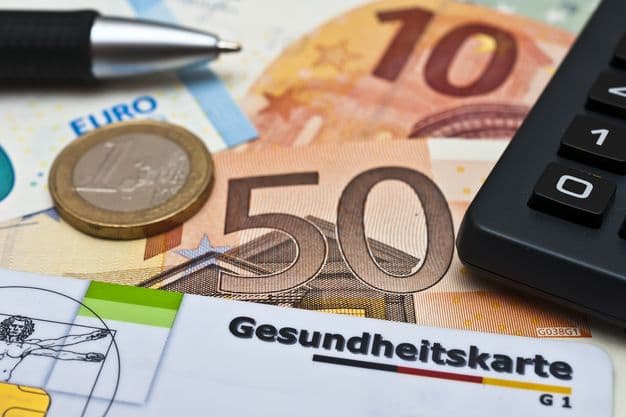 Eine Gesundheitskarte, ein Taschenrechner, ein Kugelschreiber und Geld liegen auf einem Schreibtisch.