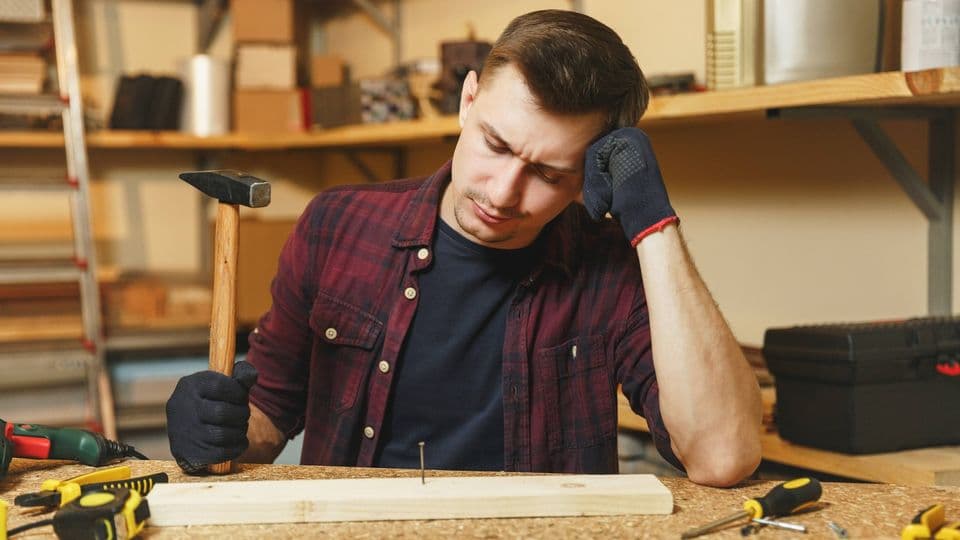 Tired upset sad caucasian young man in plaid shirt, black T-shirt, gloves hammering nails with hammer, working in carpentry workshop at wooden table place with piece of wood, different tools