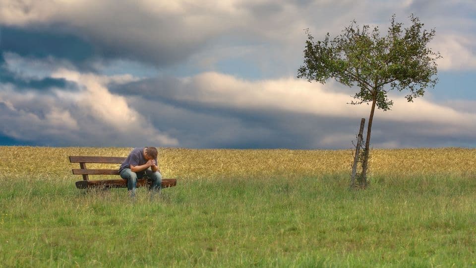 Ein Mann sitzt verzweifelt und alleine auf einer Bank, die mitten in der Natur steht.