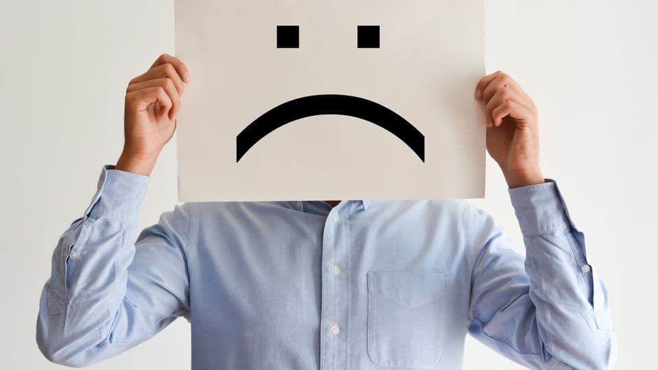 Eine Person hält sich ein unzufriedenes Gesicht auf einem Pappschild vor den Kopf.