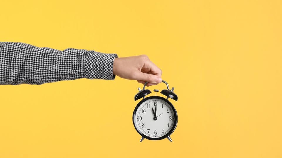 eine junge Frau hält einen Wecker vor einer gelben Wand.