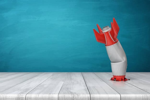 3d rendering of a red and silver realistic model of a retro rocket stands crashed into a wooden desk on a blue background. Failed launch. Technological progress. Technical mistake.