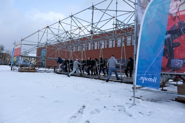 Eine Hollywood-Schaukel aus Gerüstmaterialien steht im Schnee. Einige Menschen geben der Schaukel Anschwung.