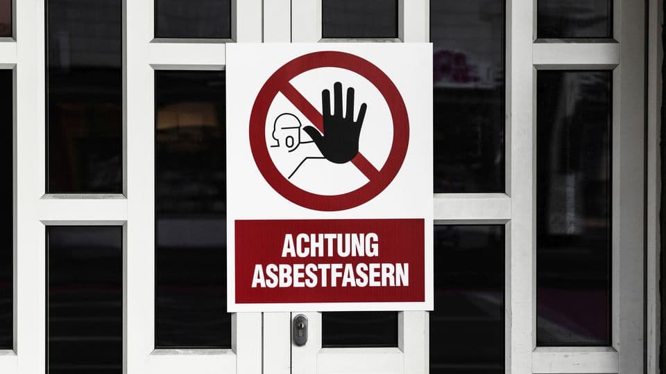 Weiße Haustür auf der ein Warnschild angebracht ist. Dort steht: "Achtung Asbestfasern".