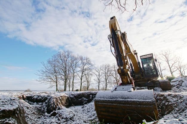 Baustelle im Winter mit Bagger, der von Schnee bedeckt ist.